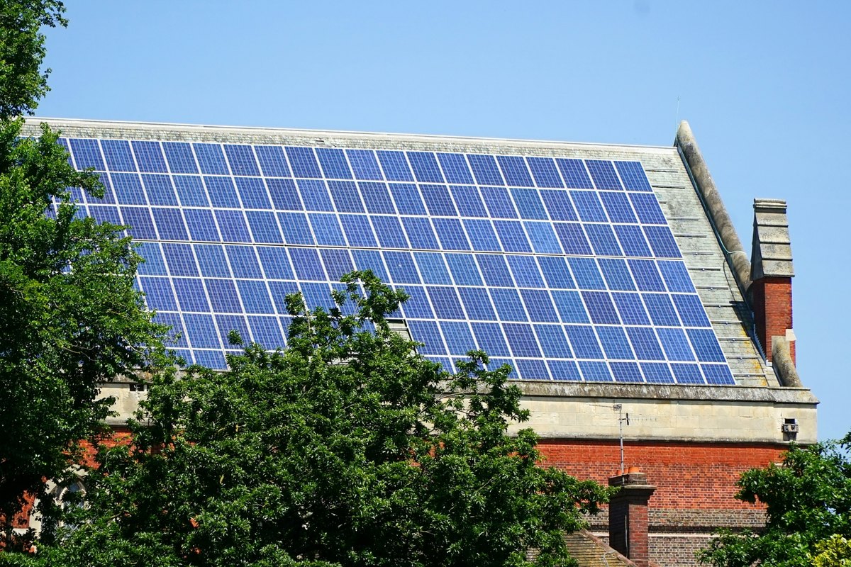 Rows of blue solar panels covering a pitched roof on a brick building with trees in front