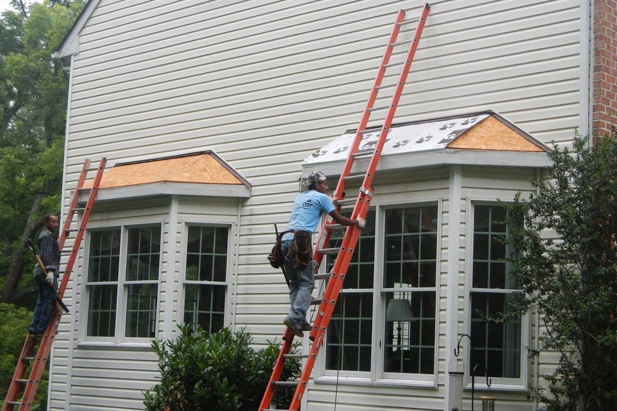 Two roofers on orange ladders working on bay window roofs on a cream vinyl-sided house