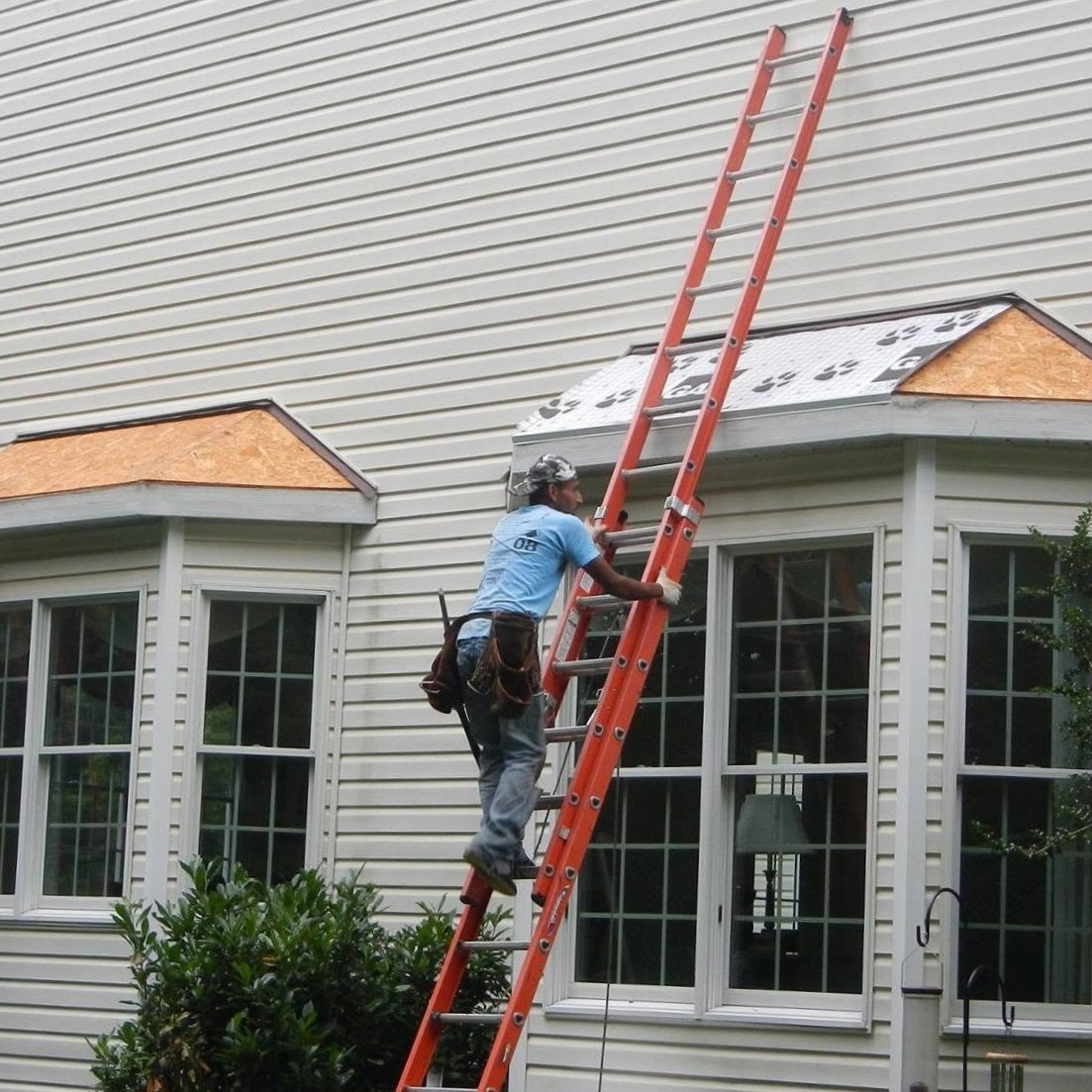 Two roofers on orange ladders working on bay window roofs on a cream vinyl-sided house