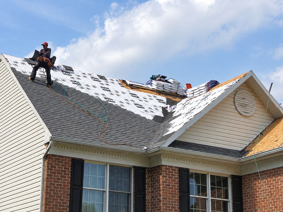 Roofer in a red cap and dark clothes sitting on the roof of a beige vinyl-sided house with a brick front and black shutters