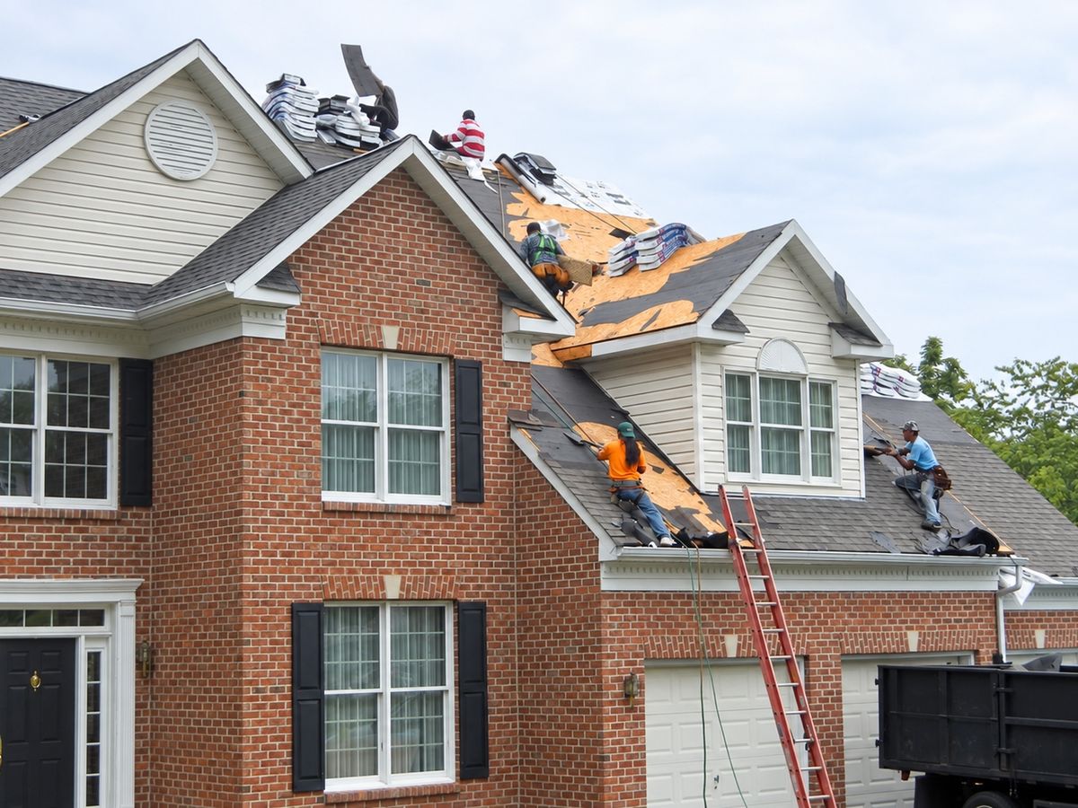 Large brick colonial with black shutters during a tear-off, four roofers working on a roof stripped to plywood
