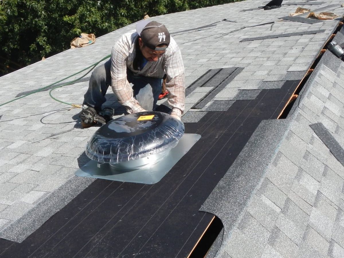 A roofer in a cap kneels on a gray shingle roof setting a plastic-wrapped solar attic fan on its metal flange