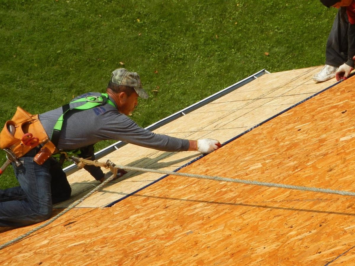 Close view of two roofers kneeling in safety harnesses on a bright orange OSB plywood roof deck with ropes