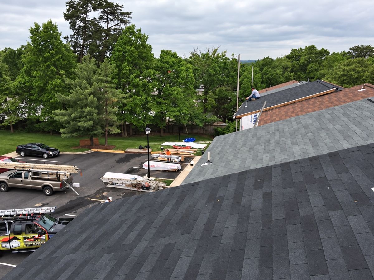 Drone view from a gray and black shingled roof ridge over an apartment parking lot: a roofer in a white shirt