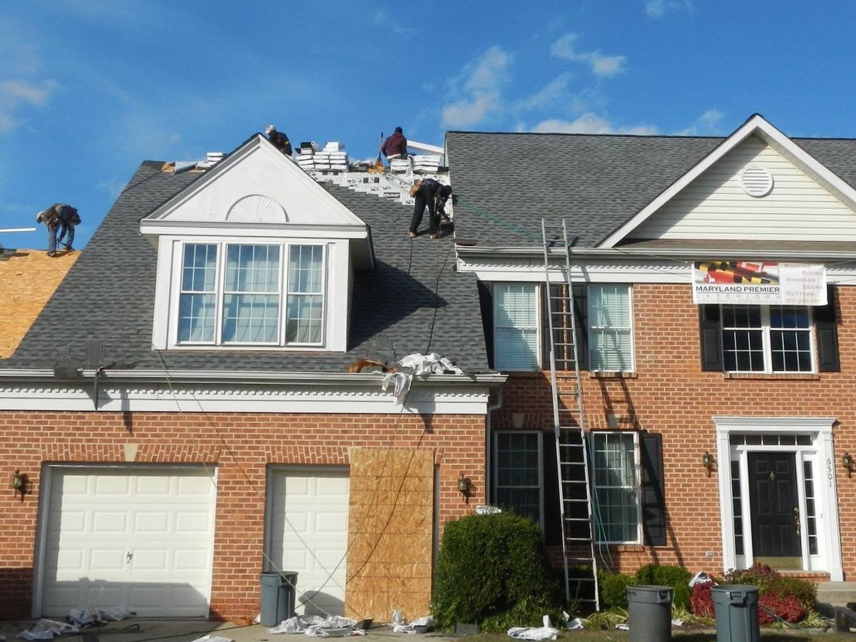 Front of a red brick colonial with a three-car garage mid re-roof under a blue sky
