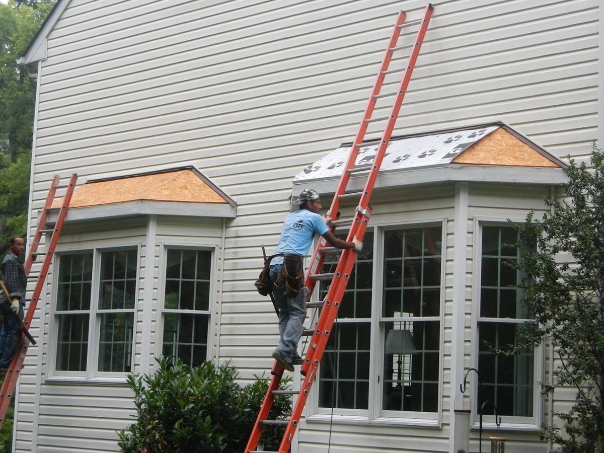 Two roofers on orange fiberglass ladders working on two small bay-window roofs on the side of a cream vinyl-sided house