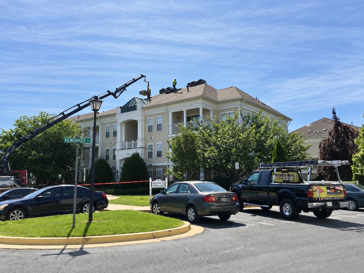 Three-story cream apartment building with a hip roof where a roofer on the ridge receives a shingle pallet