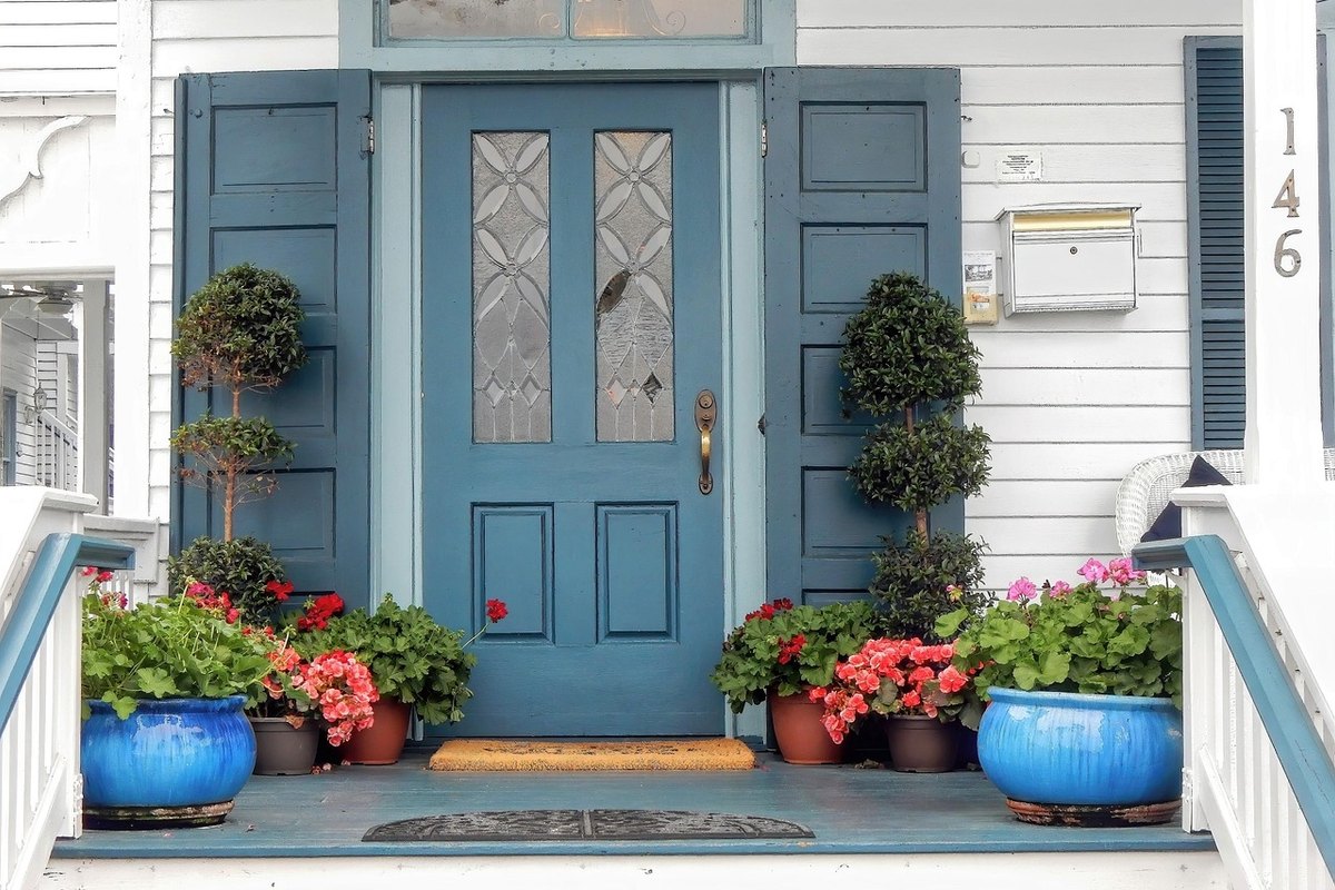 Blue front door with leaded glass panels on a white clapboard porch with potted geraniums