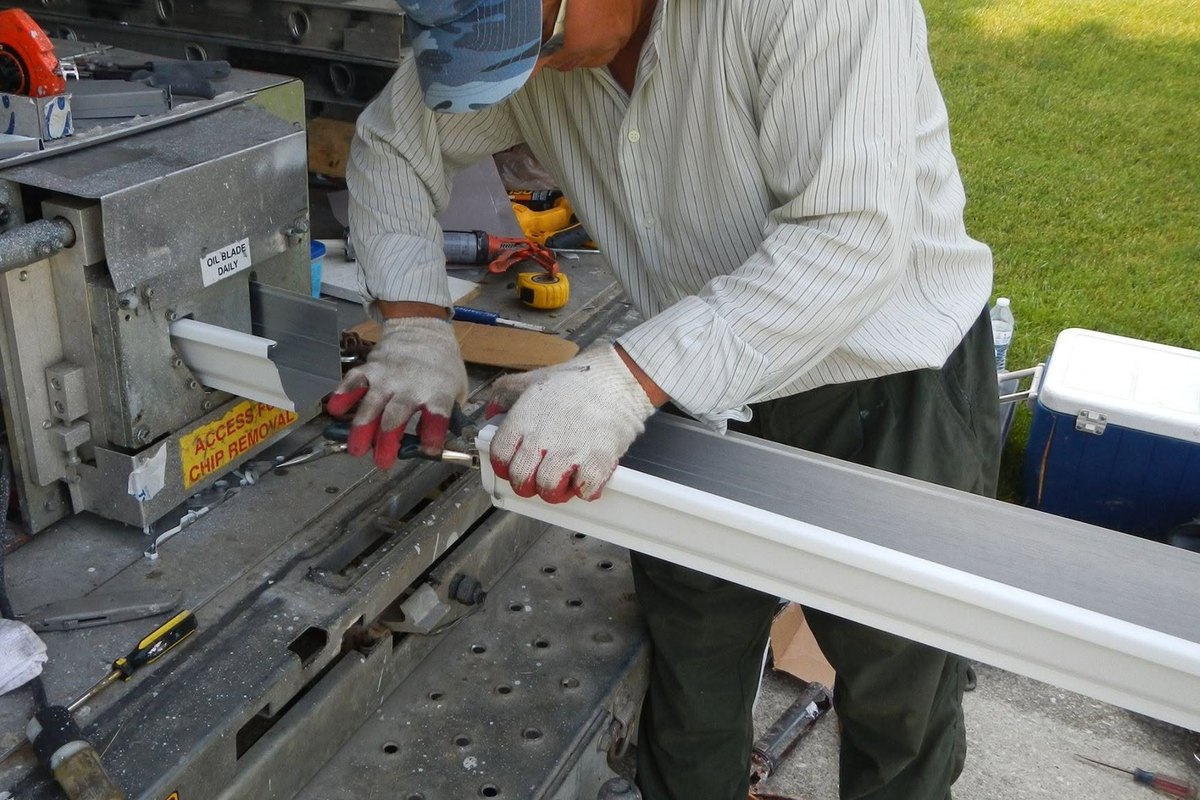 Gloved hands guiding a white seamless gutter section out of a gutter-forming machine in a work truck