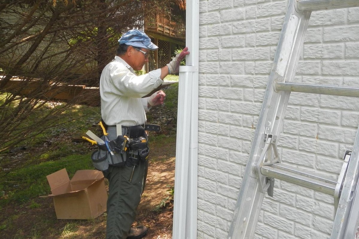 Worker in a camouflage cap installing a white downspout on a vinyl wall beside an aluminum ladder