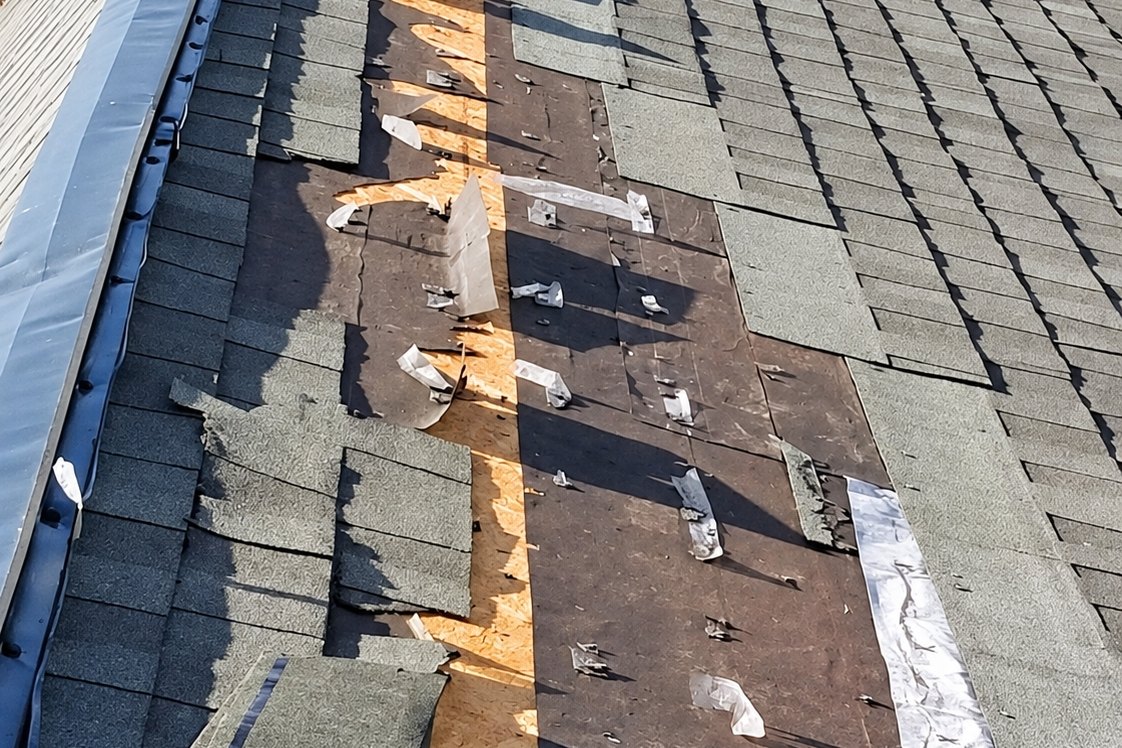 Steep asphalt shingle roof with a strip of shingles torn away down to the underlayment and plywood
