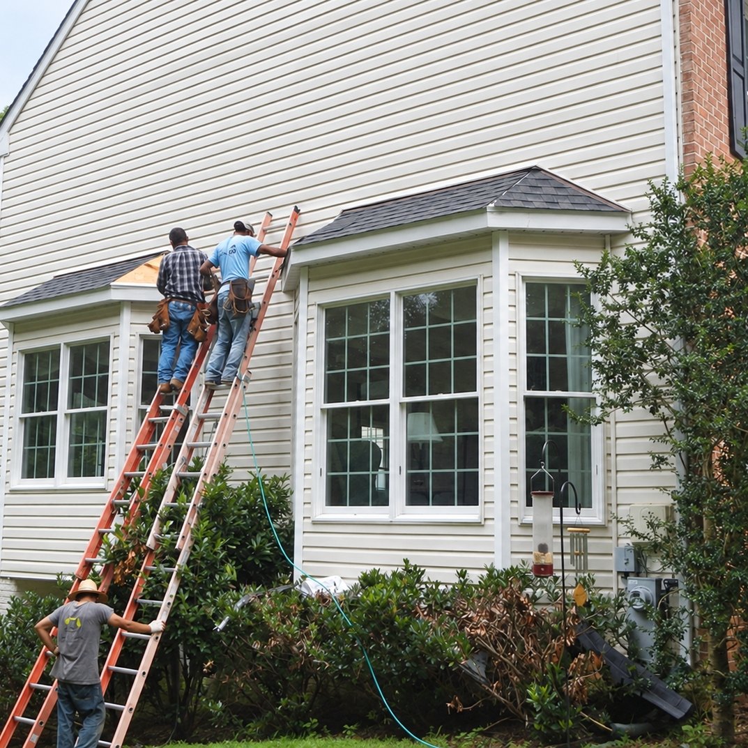 Roofers on ladders at the bay window roofs on the side of a cream vinyl-sided house