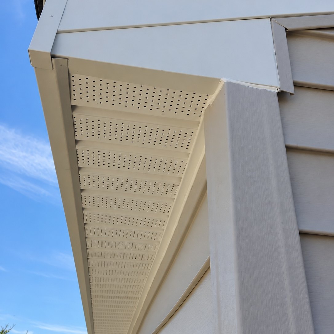 Upward view of white vinyl soffit panels, fascia and a wide corner post on a beige vinyl-sided house