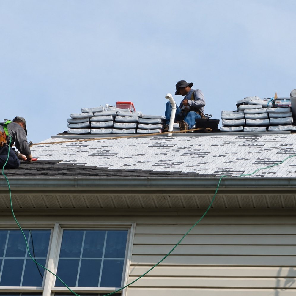 Close view from below of two roofers on the eave of a house: one kneeling and nailing dark shingles on the left